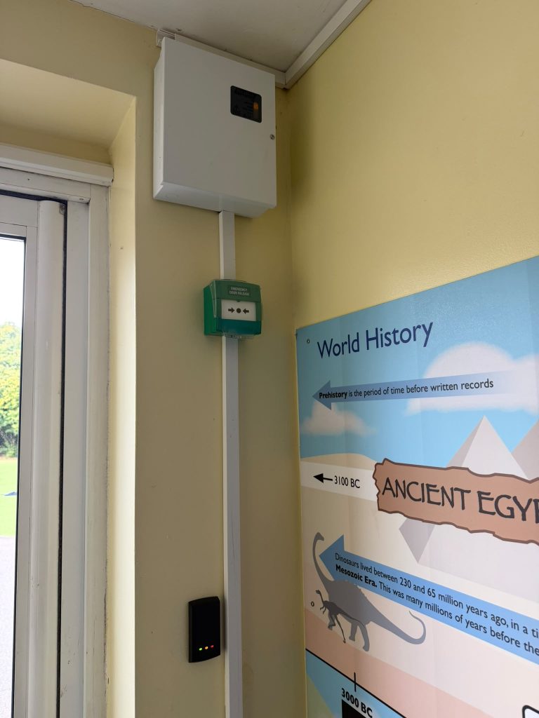 Door controller, green emergency door release and access reader beside an exit door at a primary school in Gillingham, Kent